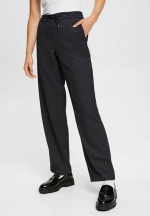 Vrouw die zwarte pantalon met krijtstreep en trekkoord draagt, zwarte chunky loafers en witte sokken, staande met één hand in de zak.