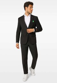 Muž v čiernom obleku s bielou košeľou, zelenou vreckovkou a bielymi teniskami stojí, pozerá na stranu na jednoduchom pozadí.