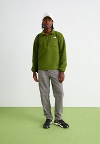 Grønn fleecegenser med kvart glidelås, frontlomme, kombinert med lysegrå jeans og svarte joggesko. Hvitt caps med tekst.