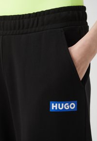 Čierne tepláky s textúrovaným materiálom, elastickým pásom, bočnou vreckom a modrou nášivkou s nápisom "HUGO."