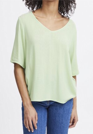Frau trägt lockeres, hellgrünes V-Ausschnitt-Kurzarmshirt und blaue Jeans, mit einer Hand in der Tasche, vor einem schlichten Hintergrund.