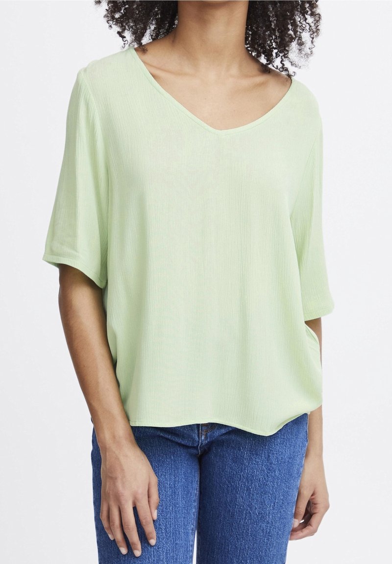 Femme portant un haut ample vert clair à col en V et manches courtes et un jean bleu, une main dans la poche, sur fond uni.
