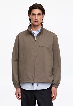 Homme portant une veste zippée marron par-dessus une chemise bleu clair et un pantalon foncé, debout devant un fond blanc uni.
