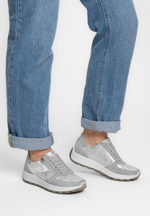 Henkilö, jolla on ylhäältä käärityt siniset farkut ja vaaleanharmaat nauhalliset tennarit, joissa on paksut valkoiset pohjat, seisoo yksinkertaista taustaa vasten.