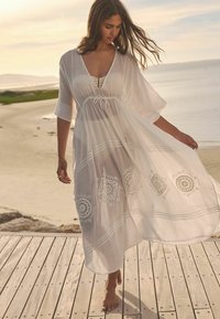 Witte, semi-doorzichtige kaftan met brede mouwen, een strik rond de taille en decoratieve borduursels langs de zoom. Een licht, vloeiend ontwerp dat ideaal is voor warm weer.