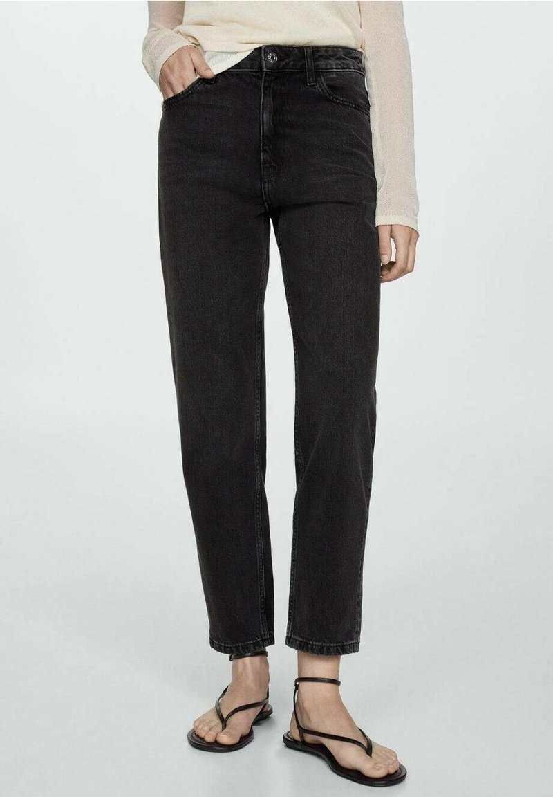 Femme portant un jean noir taille haute à jambes droites, un haut beige à manches longues et des sandales plates noires à lanières, debout avec une main dans la poche.