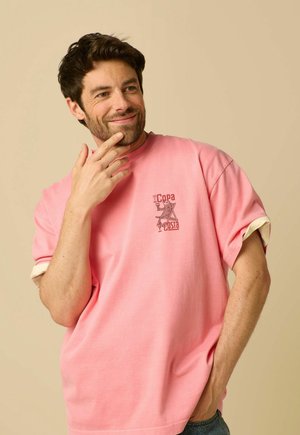 Homme souriant aux cheveux foncés portant un t-shirt rose avec un petit graphique et du texte sur la poitrine, main touchant le menton, fond beige.
