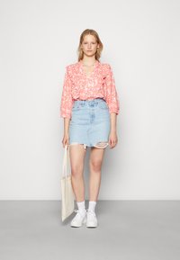 Blusa floral vermelha com detalhes em babados, combinada com uma saia de ganga azul clara com bainha desfiada e rasgos. A modelo segura uma mala tote bege.