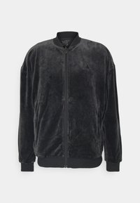 Svart velour bomberjacka med dragkedja framtill, ribbade ärmslut och nederkant, samt en broderad logotyp på vänster bröst. Mjuk textur och avslappnad passform.