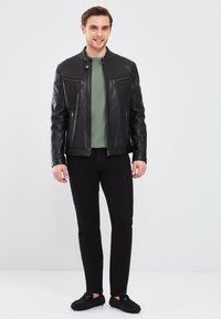 Zwarte leren jas met horizontale stiksels en twee ritszakken aan de voorkant, gecombineerd met een groene shirt en zwarte slim-fit broek.