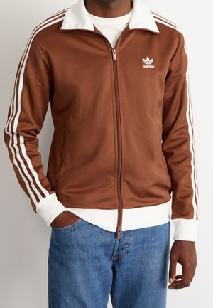Mand iført en brun Adidas lynlåsjakke med hvide striber på ærmerne og hvid krave, kombineret med blå jeans.