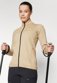 Beige zip-up jakke med et teksturert mønster, svart glidelås og oransje detaljer. Lange ermer med tommelhull; brukt med turstaver.
