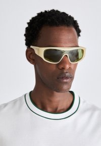 Sunglasses with a translucent yellow frame and green-tinted lenses. Angular design with wide temples, worn with a white shirt featuring green accents.