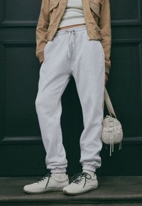 Ljusgråa bomulls-sweatpants med elastiska muddar, kombinerade med beige sneakers med svarta skosnören. Ljusbrun kort jacka och handväska.