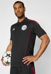Sort polo skjorte med røde sidelister og et cirkulært logo på brystet. Materialet ser glat ud; kraven er traditionel i stilen.