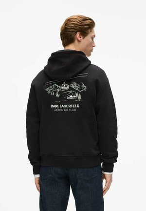 Homme de dos portant un sweat à capuche noir avec un graphisme de montagne et de remontée mécanique et le texte "Karl Lagerfeld Après Ski Club" dans le dos.