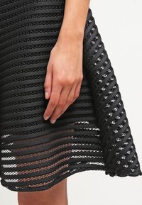 Main reposant sur le milieu de la cuisse, jupe noire avec des rayures horizontales alternées opaques et transparentes et un motif texturé.