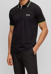 Svart polotröja med vit och grönrandig krage. Har tre knappar, broderad "BOSS"-logotyp på bröstet, och mjuk textur.