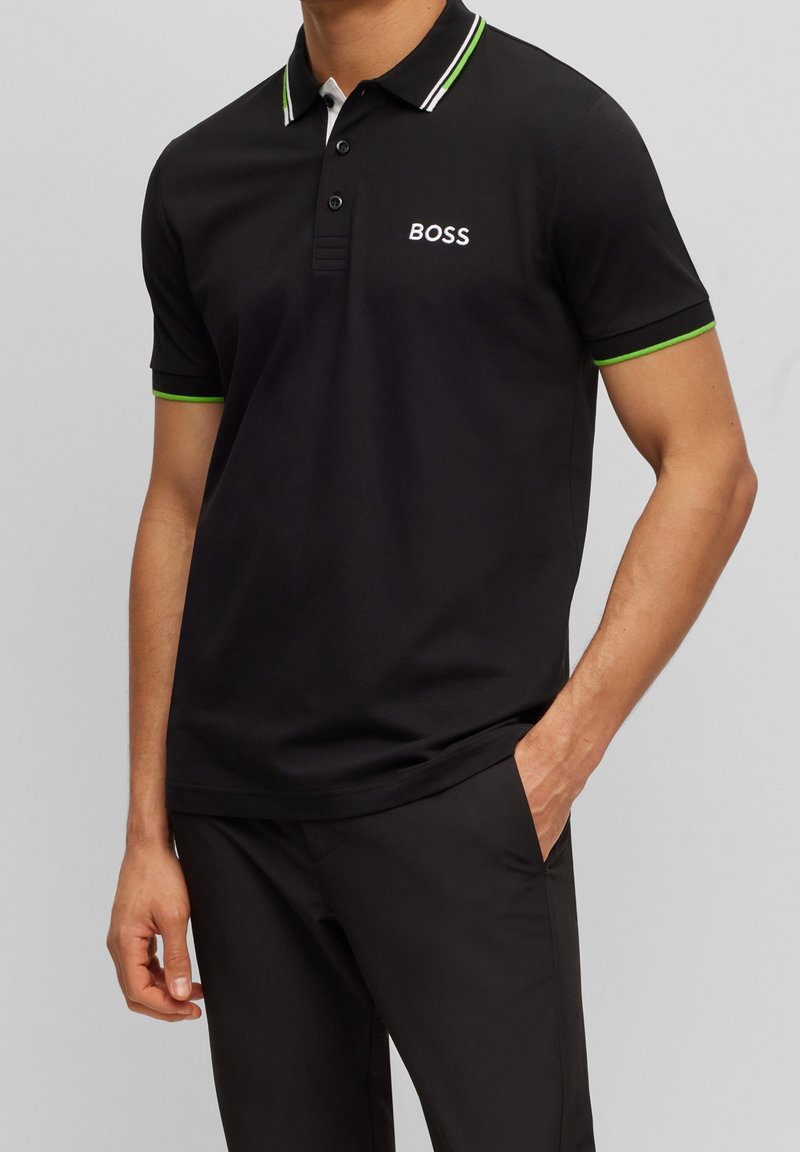 Svart polotröja med vit och grönrandig krage. Har tre knappar, broderad "BOSS"-logotyp på bröstet, och mjuk textur.