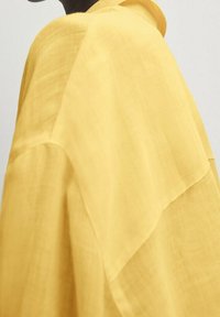 Blouse jaune, légère, avec une coupe ample, présentant une texture douce et un encolure large. Le tissu a un éclat subtil et est semi-transparent.