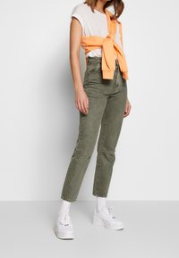 Jeans de talle alto verde oliva, camiseta a rayas blanca y gris, sudadera naranja atada a la cintura, zapatillas blancas con suelas gruesas.