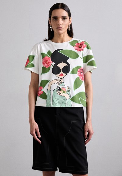 Hvit avkortet t-skjorte med blomster- og illustrasjonsdesign, som viser en figur som holder en tekopp, kombinert med et svart skjørt med lommer.