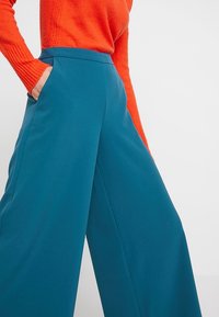 Vidbyxa i turkos tyg, hög midja med framfickor, tillsammans med en orange strukturerad tröja. Tydliga linjer och avslappnad stil.