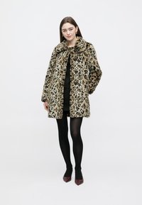Casaco de pelo falso com estampa de leopardo, gola larga e frente aberta, com tons de castanho e preto, combinado com um vestido preto e collants pretos.