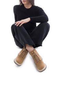Botas de ante color beige con cordones, suela de goma, ojales metálicos y una entresuela blanca en contraste, combinadas con pantalones de pierna ancha negros.