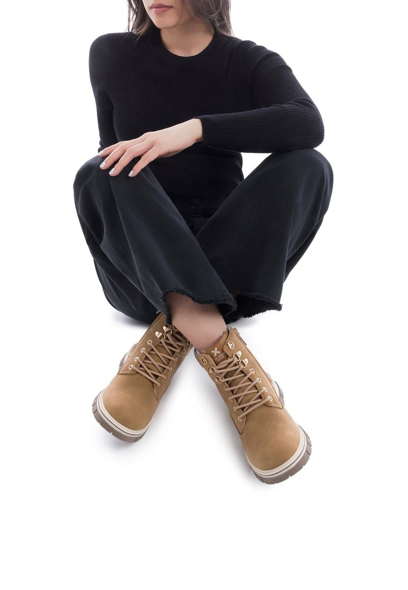 Botas de ante color beige con cordones, suela de goma, ojales metálicos y una entresuela blanca en contraste, combinadas con pantalones de pierna ancha negros.