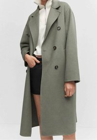 Manteau croisé vert olive en mélange de laine, avec de gros boutons noirs, de larges revers et une coupe décontractée, porté sur un haut à col montant et une jupe.