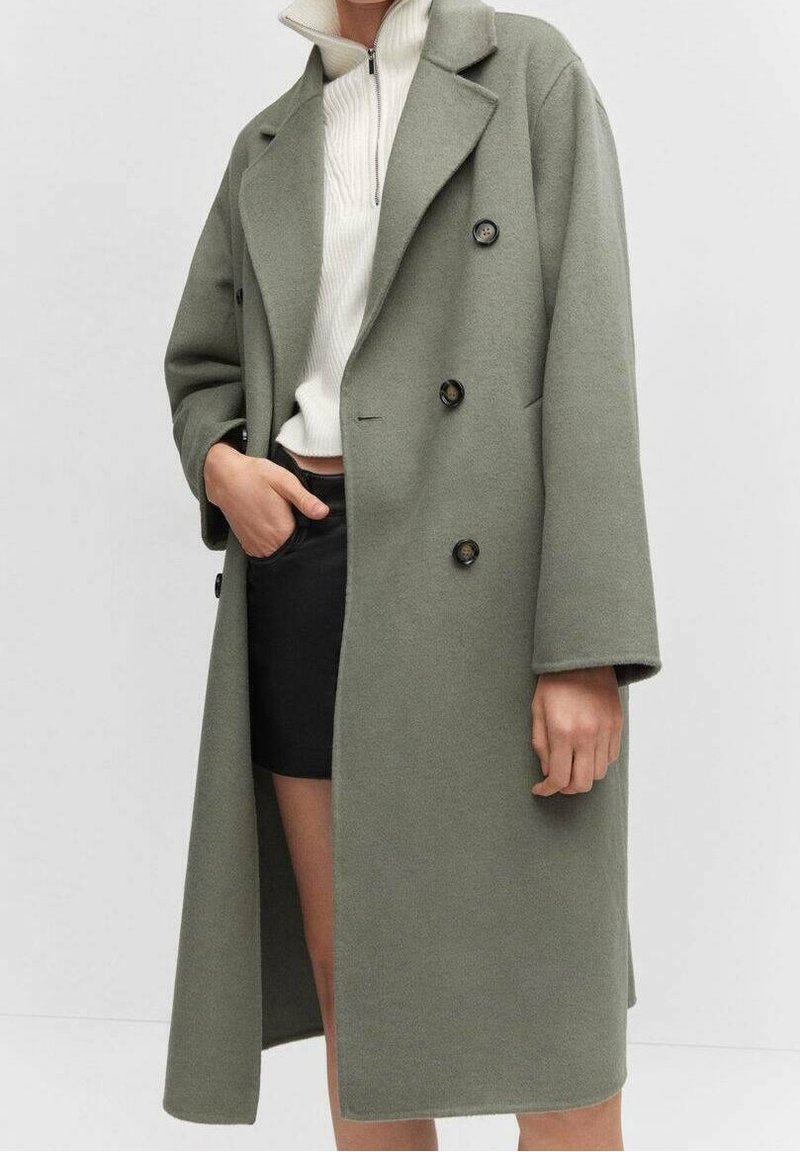 Manteau croisé vert olive en mélange de laine, avec de gros boutons noirs, de larges revers et une coupe décontractée, porté sur un haut à col montant et une jupe.