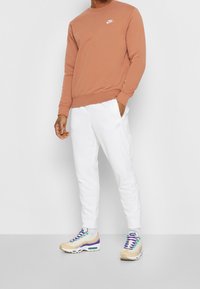 Nike huppari terrakottavärissä, jossa on pyöreä kaula-aukko ja valkoinen logo. Yhdistetty valkoisiin collegehousuihin, joissa on sivustripit ja beiget kengät.