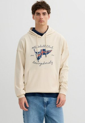 Joven con sudadera con capucha beige con el texto "Whitehall Club Rowing Society" y una bandera gráfica, combinado con jeans azules, posando frente a un fondo liso.