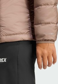 Brązowa kurtka puchowa o pikowanej fakturze, z brandingiem „ClimaWarm” na rękawie. Czarna dopasowana spodnie z białym logo.