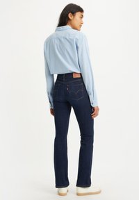 Chemise col bleu clair et jean en denim bleu foncé évasé avec une étiquette en cuir sur la taille, doté de surpiqûres contrastantes. Baskets blanches.