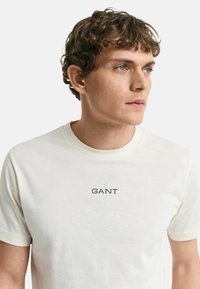 Camiseta de algodón blanca de manga corta con cuello redondo y un logo en navy "GANT" bordado en el centro del pecho. Textura suave.