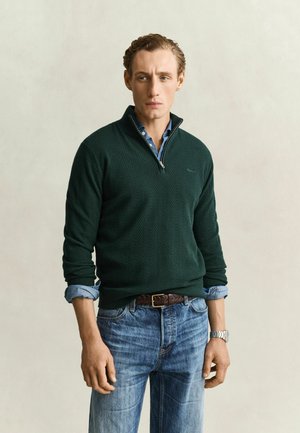 Jonge man met lichtbruin haar, draagend een donkergroene trui met ritssluiting over een blauw overhemd en blauwe spijkerbroek, staand tegen een effen lichte achtergrond.
