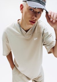 Beige mesh shirt met korte mouwen, een V-hals en een subtiele logo. Een zilveren kettinghanger en een versleten grijze pet worden gedragen.