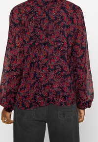 Blusa floreale navy con maniche lunghe trasparenti, caratterizzata da motivi floreali rossi, rosa e blu, e un orlo arrotondato sul retro.