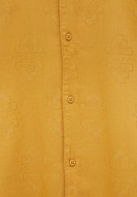 Košile na knoflíky v hořčicově žluté barvě s texturovaným vzorem. Má čtyři shodné knoflíky vpředu. Hladký povrch látky.
