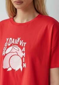 Rode katoenen t-shirt met een grafisch ontwerp van fruit en de tekst "JOUW DAGELIJKSE VITAMINE" in het wit, met een relaxte pasvorm en korte mouwen.