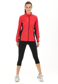 Veste à fermeture éclair rouge avec des panneaux latéraux noirs, col haut et accents réfléchissants ; associée à des leggings capri noirs et des chaussures de sport légères.