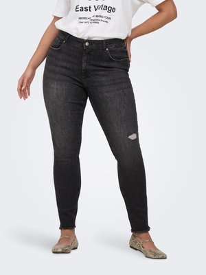 Femeie purtând blugi skinny negri rupți, tricou alb cu grafică și balerini cu imprimeu de șarpe, stând cu o mână în șold.