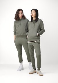 Sudaderas y pantalones de chándal en color verde oliva, fabricados en un tejido suave, con cordones, bolsillos y puños acanalados. Logo distintivo en el pecho.