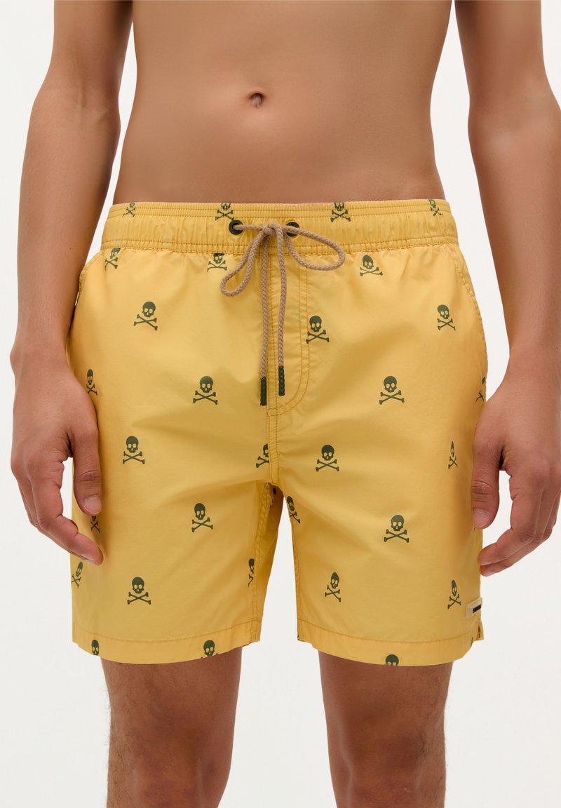 Pantaloni de baie galbeni cu un model repetat de crani și oase închise la culoare, cu un elastic la talie și închidere cu șnur.