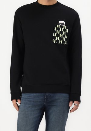 Person trägt ein schwarzes Langarmshirt mit gemusterter Brusttasche, auf der eine kleine Grafik einer Person mit weißen Haaren und Sonnenbrille zu sehen ist.