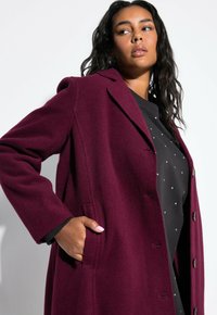 Cappotto in misto lana bordeaux con colletto a risvolto, chiusura con bottone e tasche laterali, indossato sopra un top nero con decorazioni argentate.