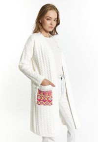 Cardigan bianco lavorato a maglia con un design a cavo testurizzato, caratterizzato da una tasca decorata con un vivace motivo nei colori rosa, giallo e viola. Maniche lunghe e apertura frontale.