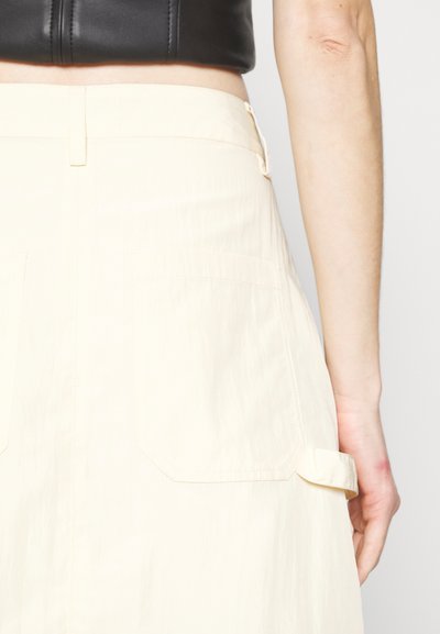 Jupe de couleur crème avec une texture lisse, dotée de deux poches arrière et d'un détail de sangle sur le côté. Se porte légèrement au-dessus de la taille.